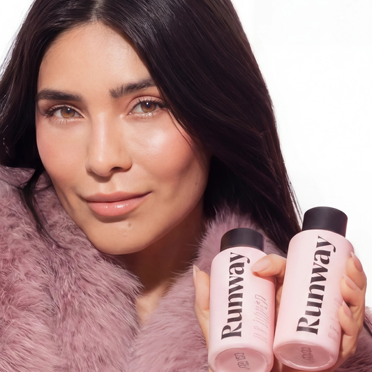 Person with long dark hair wearing mauve faux fur, holding two identical pink cosmetic bottles labeled Runway with black caps.