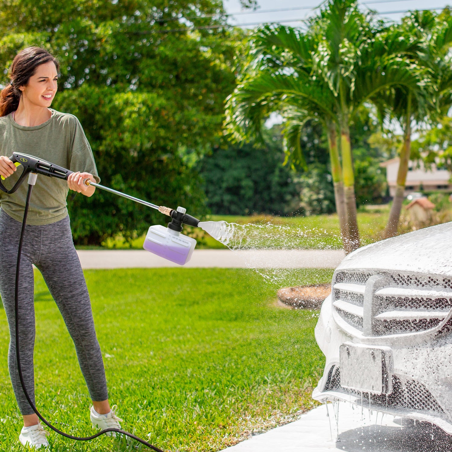 Woman using a pressure washer to clean a car outdoors on a sunny day.