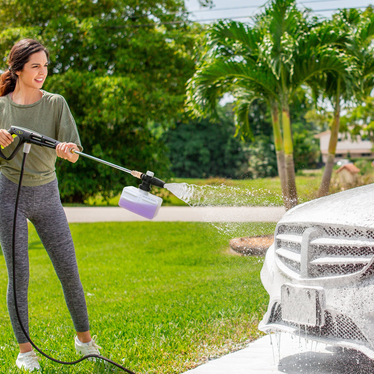 Woman using a pressure washer to clean a car outdoors on a sunny day.