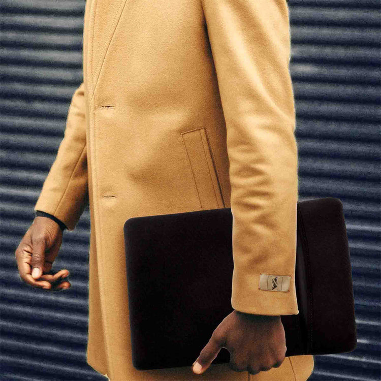 Person wearing a beige coat holding a black clutch bag against a blue corrugated metal background