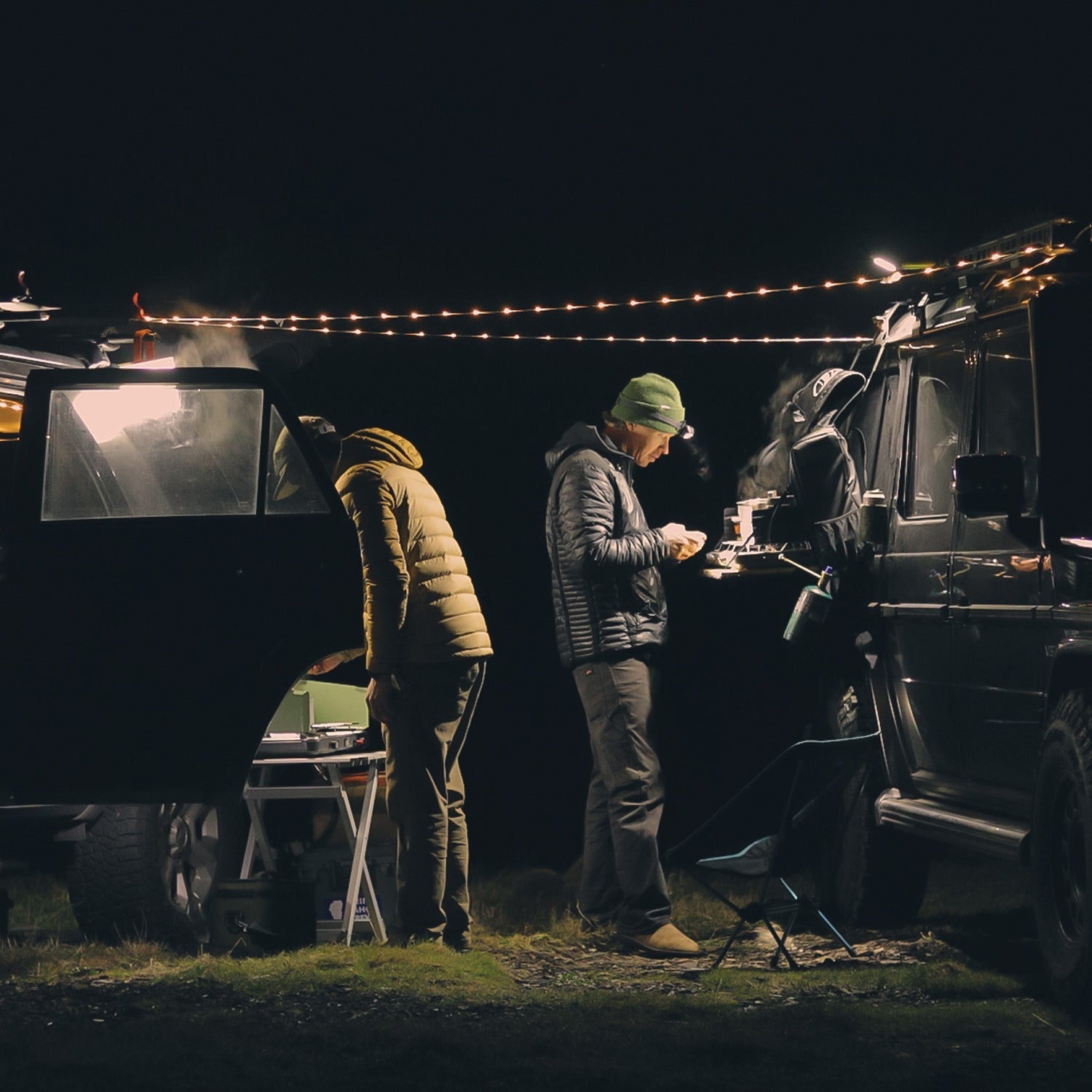 Three people gathered around a vehicle at night with string lights above.