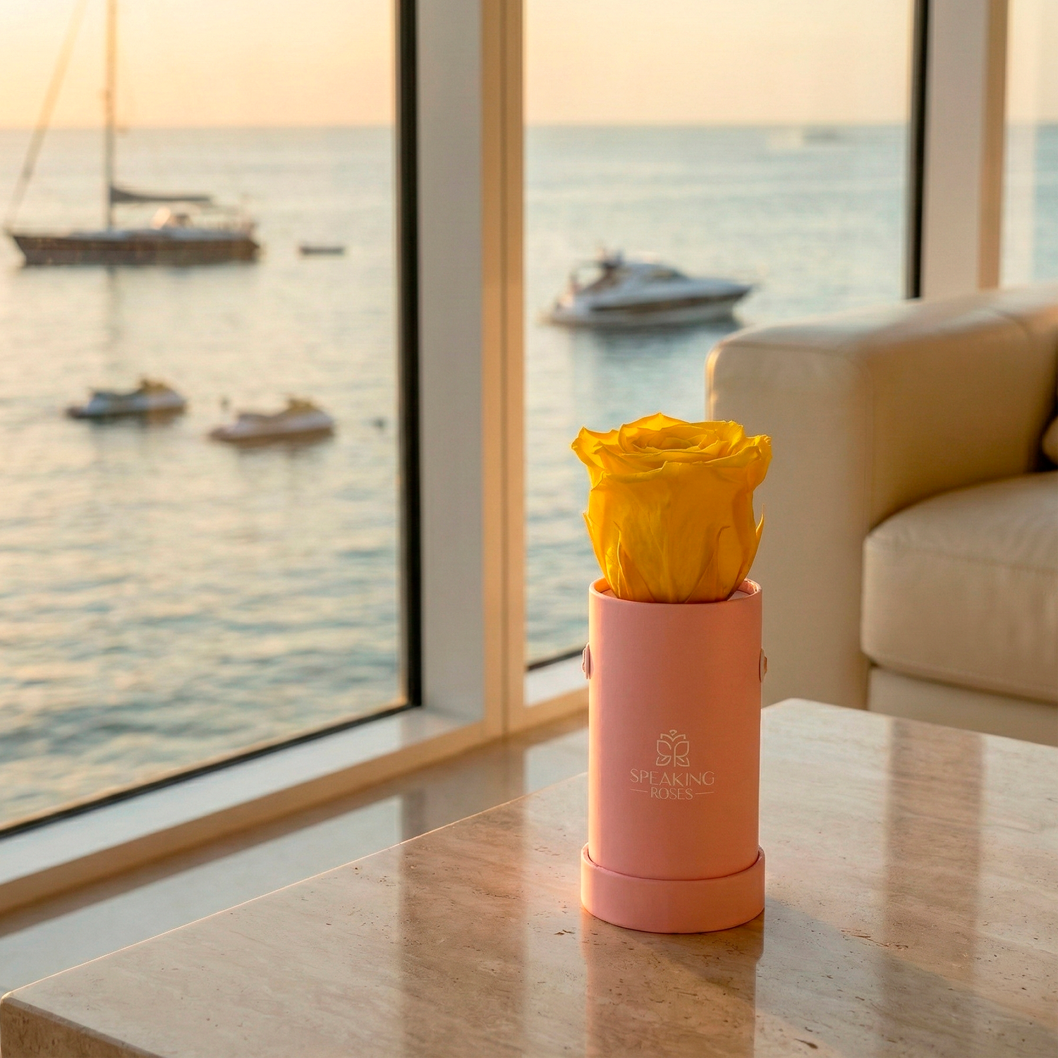 Pink vase with yellow flowers on a table by a large window with a view of the sea and boats.