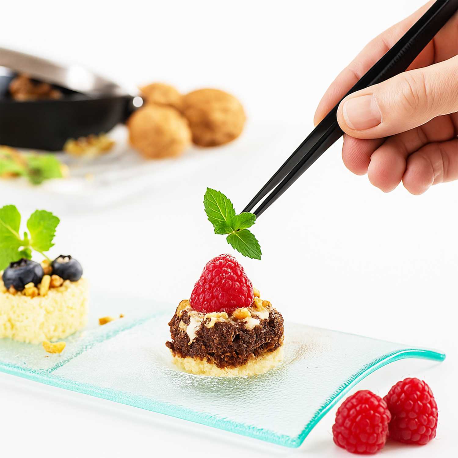 Dessert with a raspberry on a glass plate, and a hand holding tweezers placing a mint leaf on it.