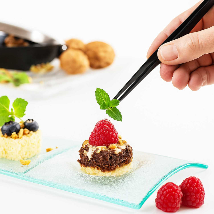 Dessert with a raspberry on a glass plate, and a hand holding tweezers placing a mint leaf on it.