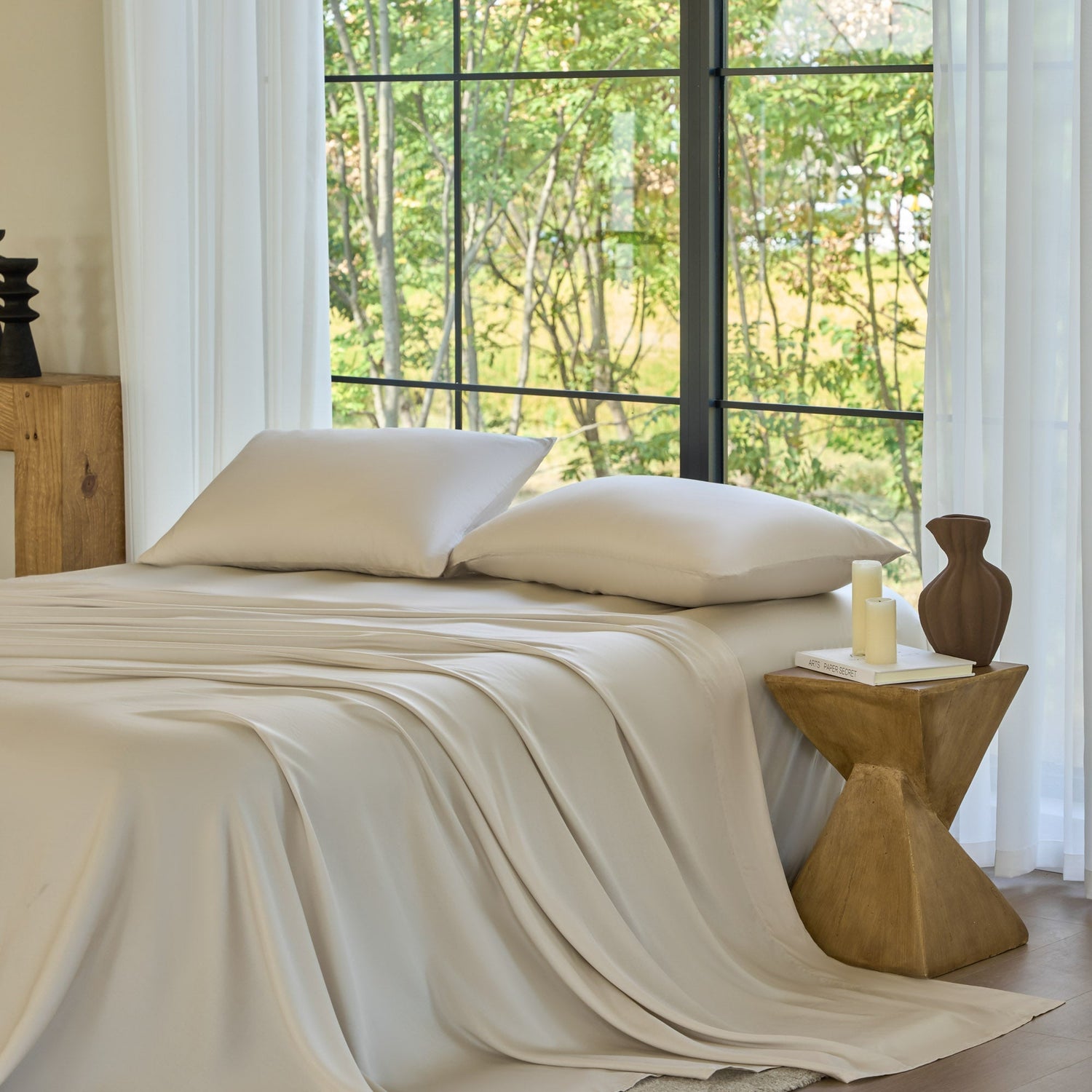 Neatly made bed with beige bedding in a room with large windows and a wooden side table.