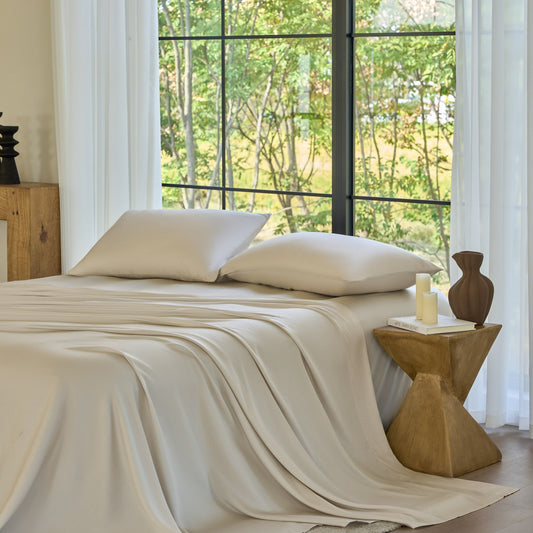 Neatly made bed with beige bedding in a room with large windows and a wooden side table.