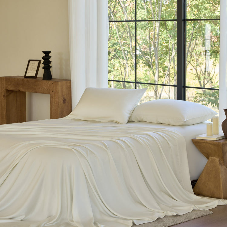 Neatly made bed with white bedding in a room with large windows and wooden furniture.