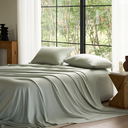 Bed with light green bedding and pillows in a room with large windows showing trees.