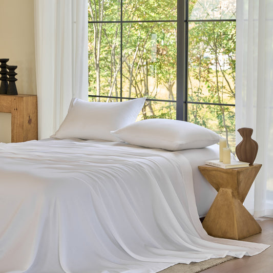 Bed with white bedding and pillows in a room with large windows and wooden side table.
