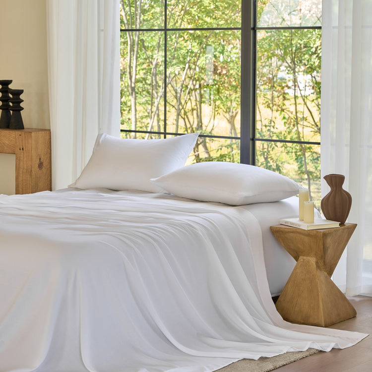 Bed with white bedding and pillows in a room with large windows and wooden side table.
