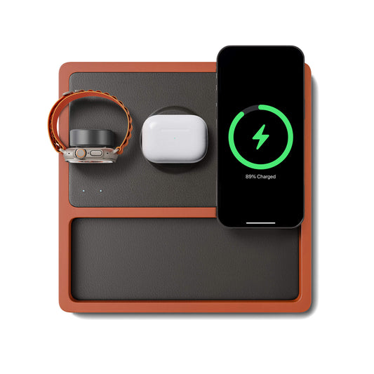 A wireless charging pad with integrated Apple Watch charger, featuring a black and oak design, with a smartphone, smartwatch, and earbuds being charged.