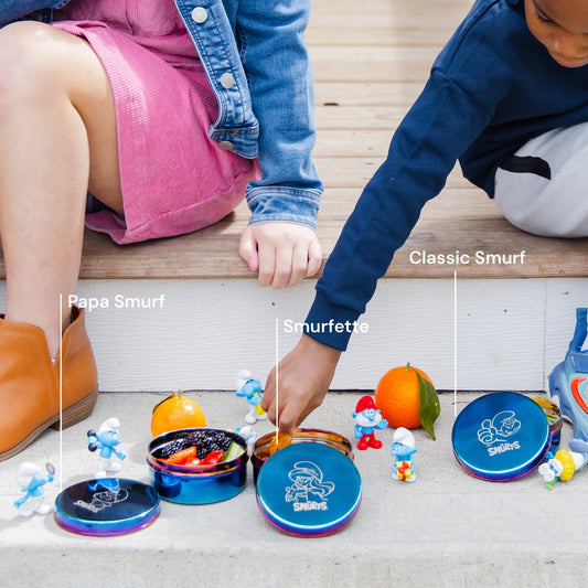 Three blue metal containers with colorful rims on a white background - each with a Smurf etched on the lid along with The Smurfs logo - Papa Smurf, Smurfette, and Classic Smurf