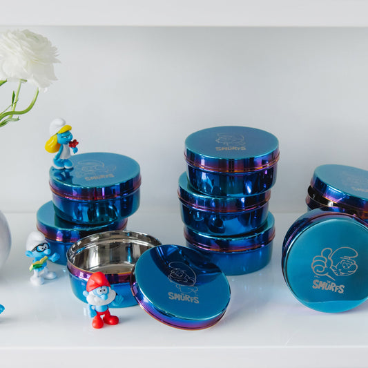 Three blue metal containers with colorful rims on a white background - each with a Smurf etched on the lid along with The Smurfs logo - Papa Smurf, Smurfette, and Classic Smurf