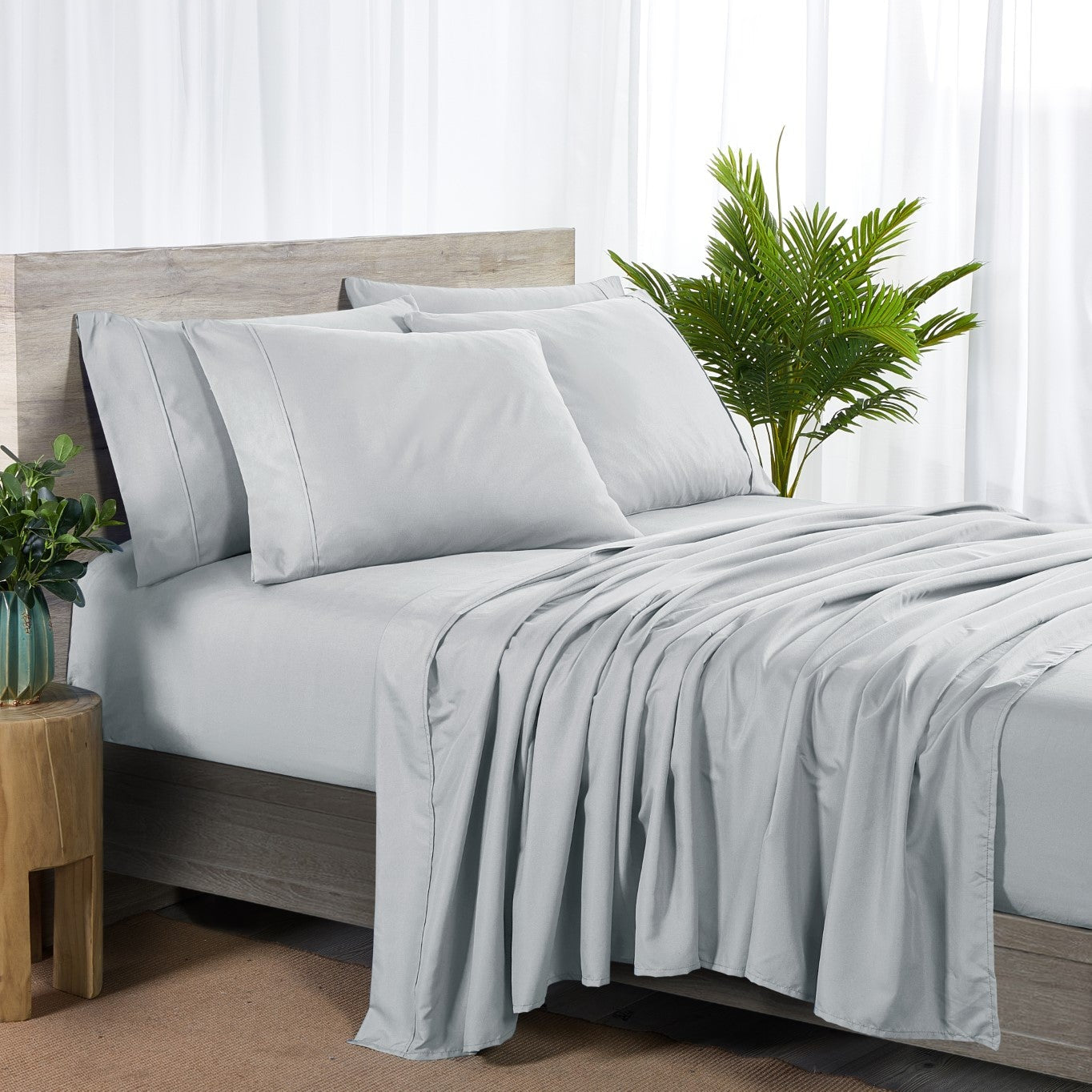 A silver 6-piece bamboo sheet set on a bed, including a fitted sheet, a flat sheet, and pillowcases, presented in a well-lit room with a plant and nightstand beside the bed.