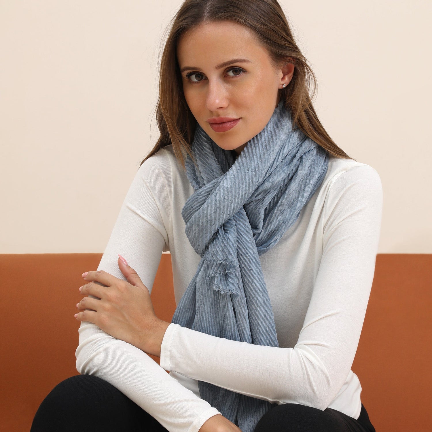 A woman wearing a lightweight blue and white striped scarf around her neck, with a white top and dark pants, sitting against a neutral background.