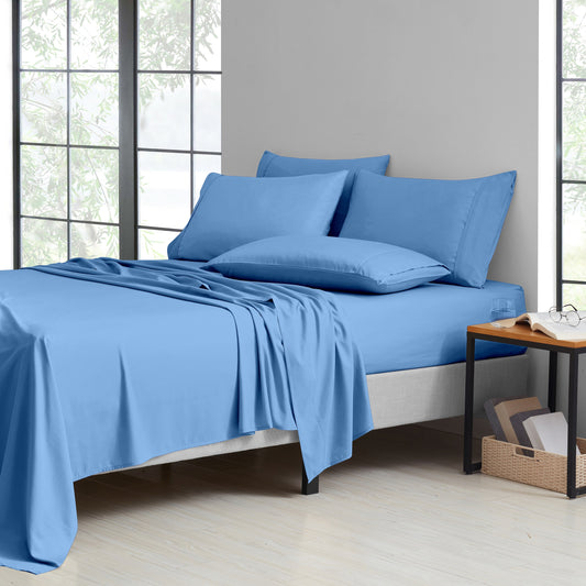 A neatly made bed with a light blue bedding set, including a fitted sheet, pillowcases, and a draped duvet, shown in a bright bedroom with large windows and a small wooden side table.