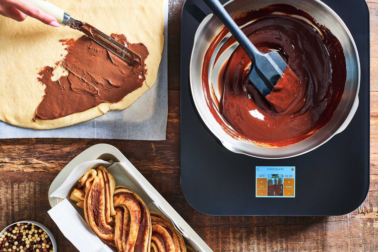 Chocolate being spread on a pastry with a spatula, next to a bowl of melted chocolate and a baked pastry.