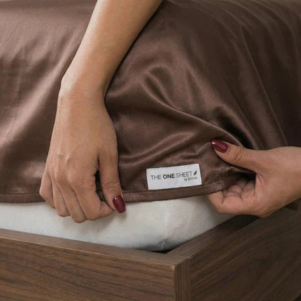 Brown satin sheet being adjusted with a visible brand label on a wooden bed frame.