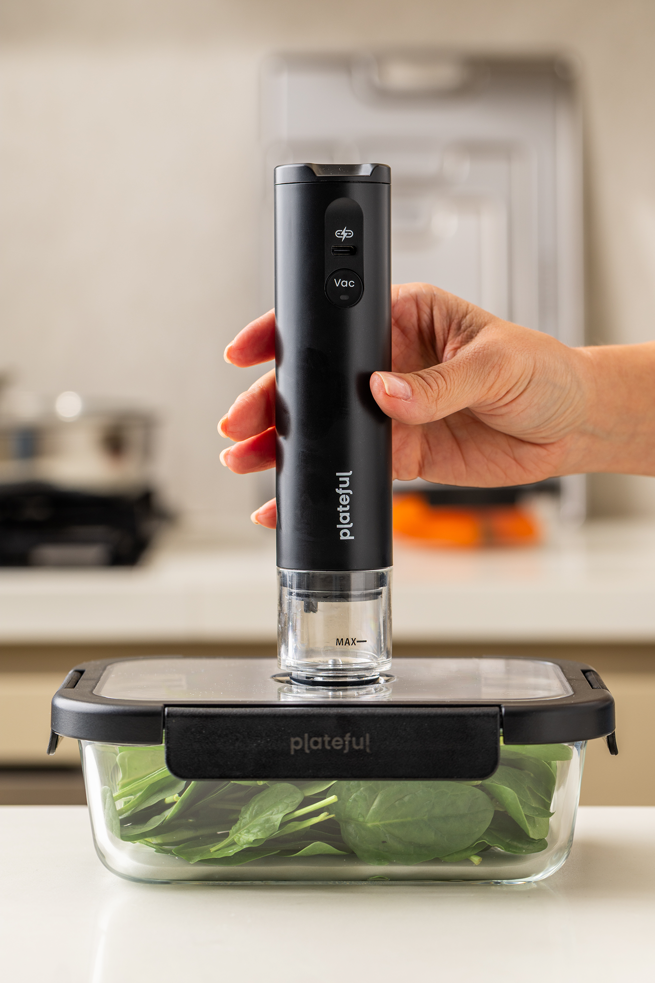 Hand holding a black plateful vacuum sealer over a container of greens on a kitchen counter.