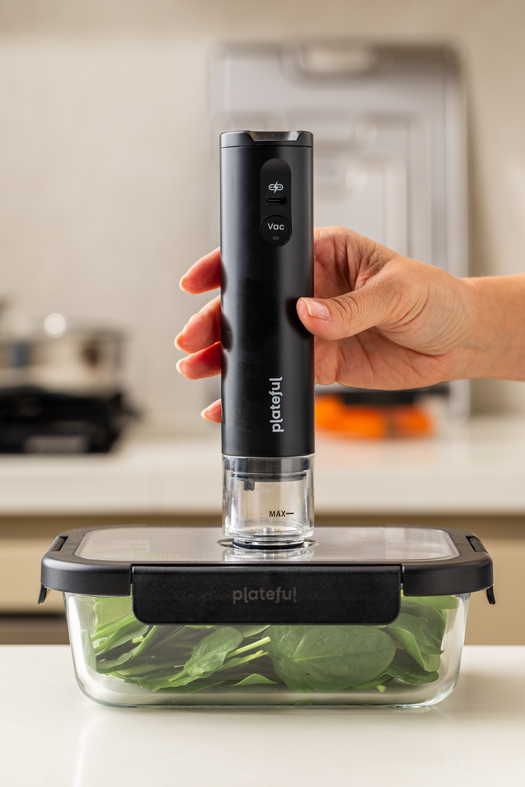 Hand holding a black plateful vacuum sealer over a container of greens on a kitchen counter.