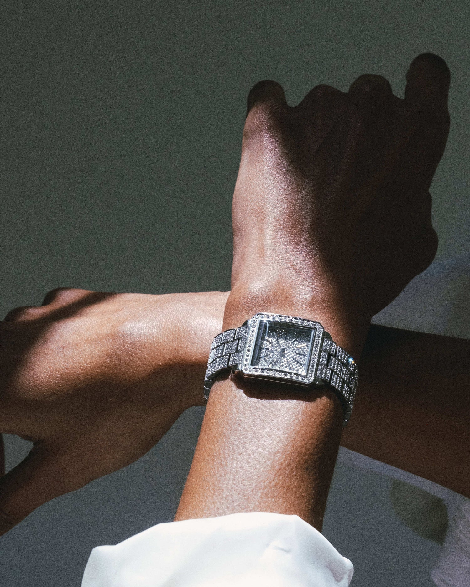 A close-up shows a wrist wearing the JBW Cristal 28 | J6386C, a square, diamond-studded stainless steel watch. Another hand is raised above against a neutral background, with dramatic lighting highlighting the watch’s exquisite details.