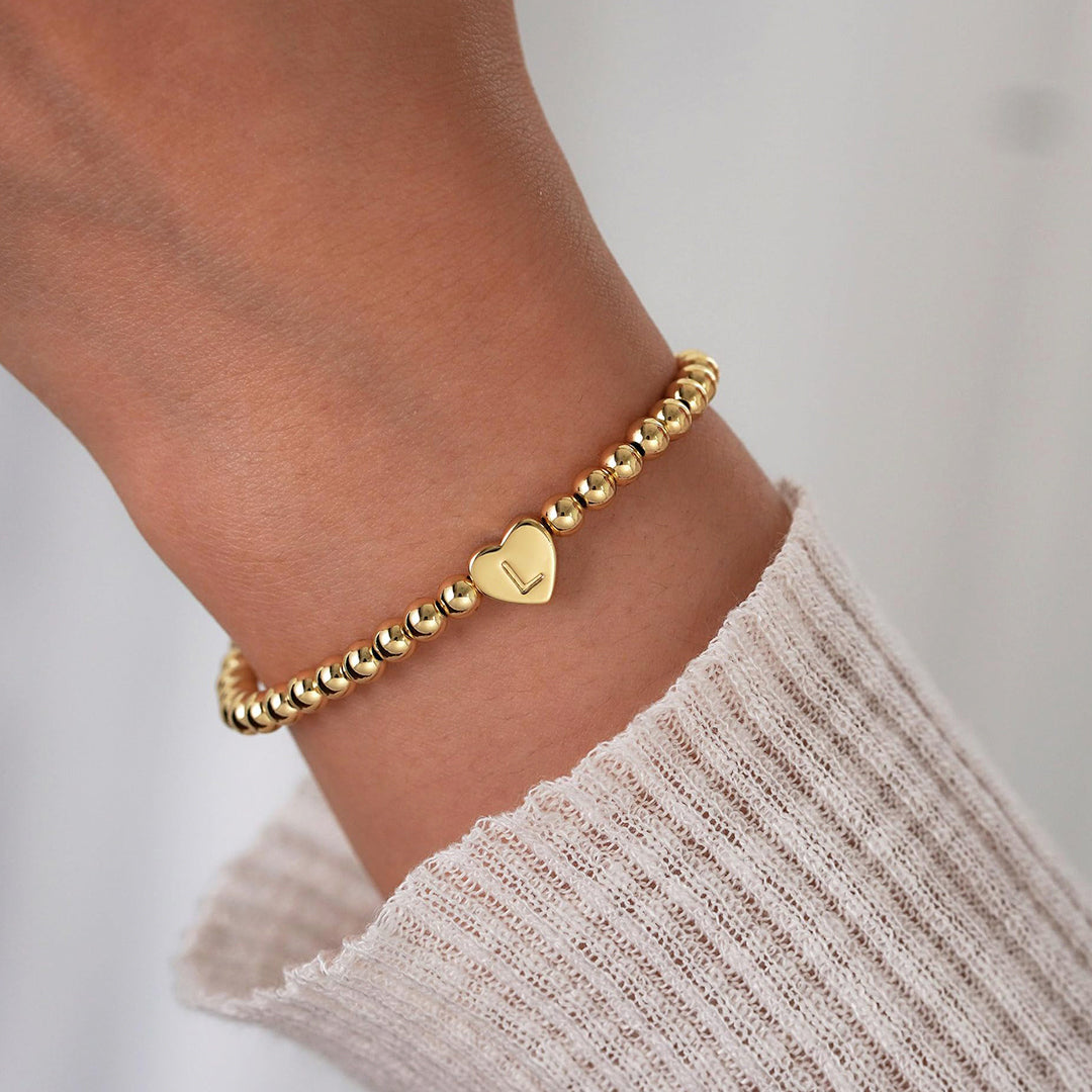 Gold beaded bracelet with a heart-shaped tag on a wrist against a neutral background