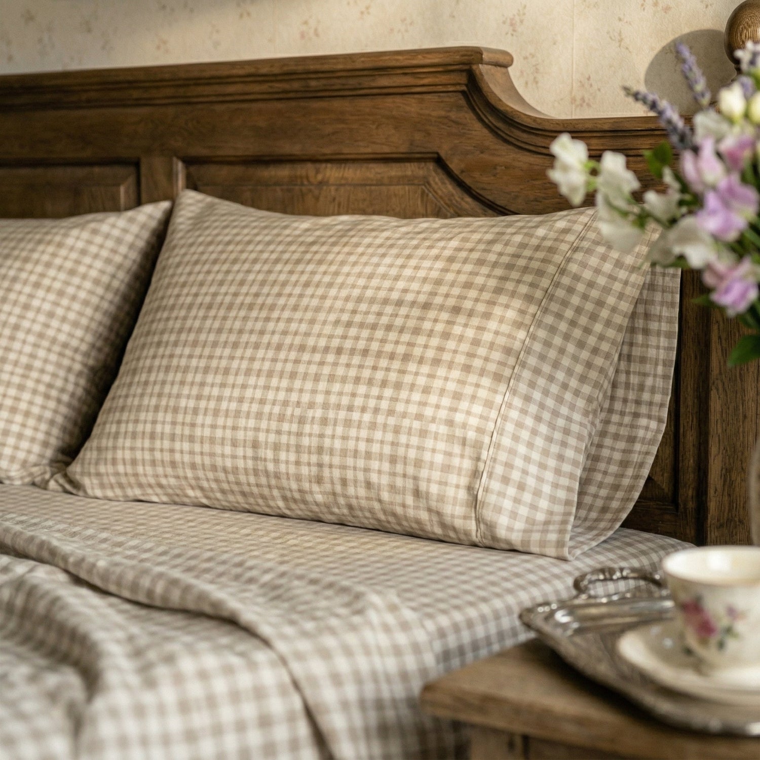 A close-up of a bed dressed in beige and brown gingham check bedding, with a dark wooden headboard, floral accents, and a bedside table in a softly styled bedroom setting.