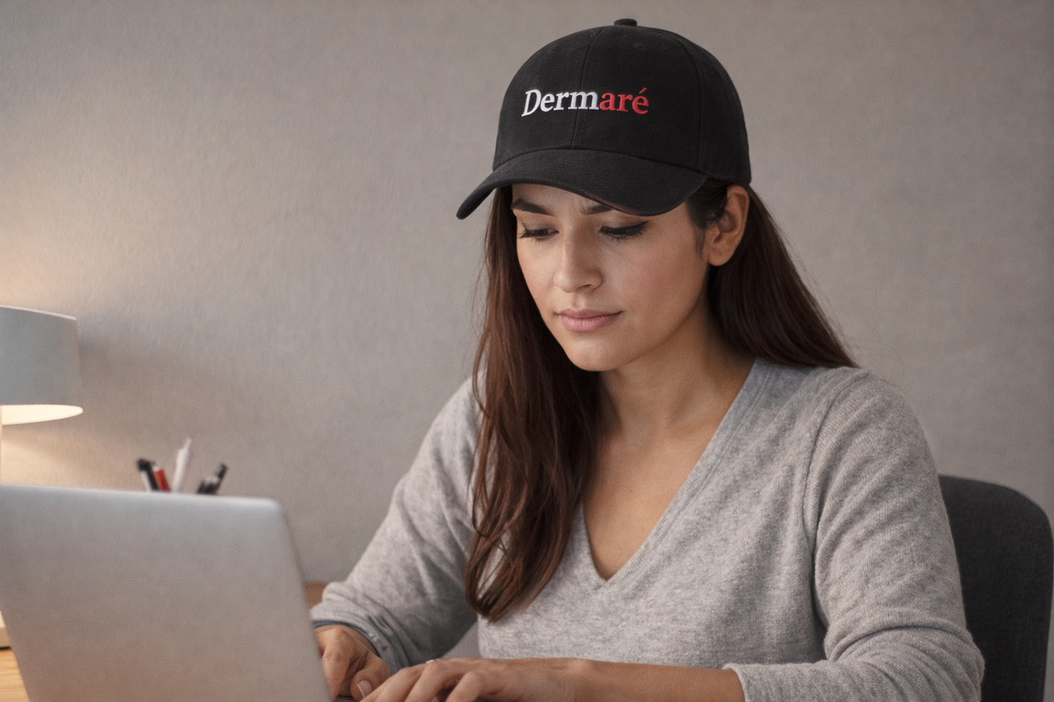 Woman wearing a black cap with 'Dermare' logo using a laptop.
