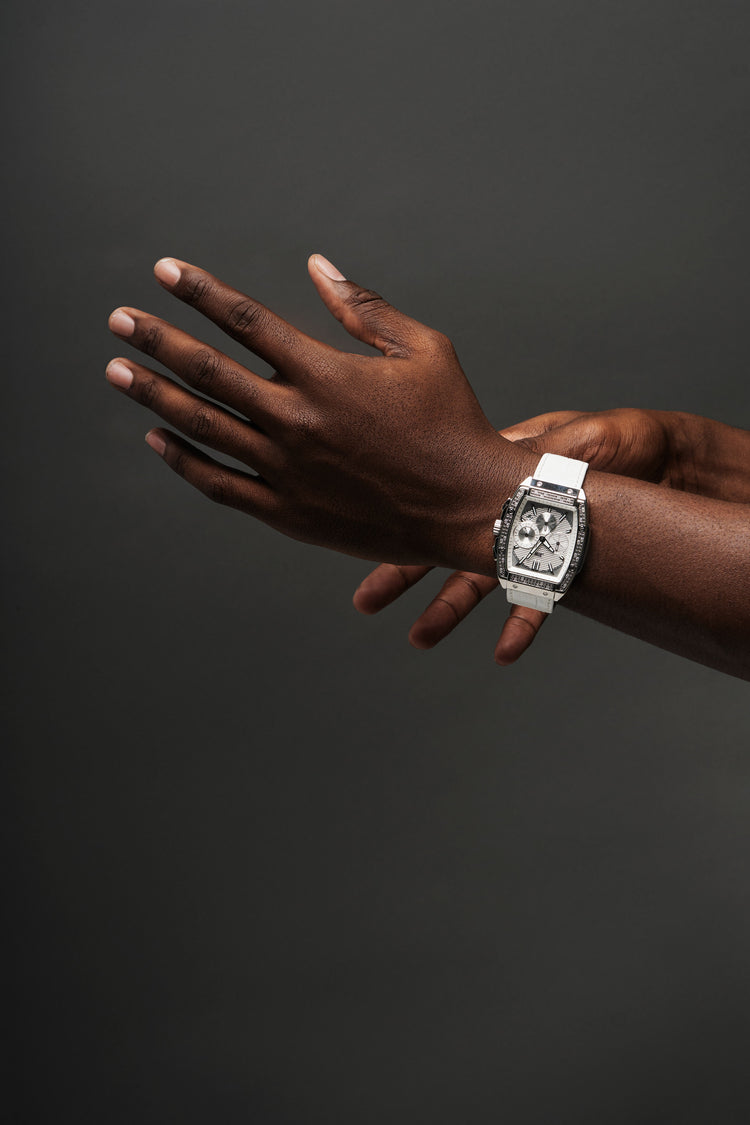 A close-up of a person's hands and forearms wearing a white rectangular watch with a silver-toned bezel and dial details, set against a dark gray studio background.