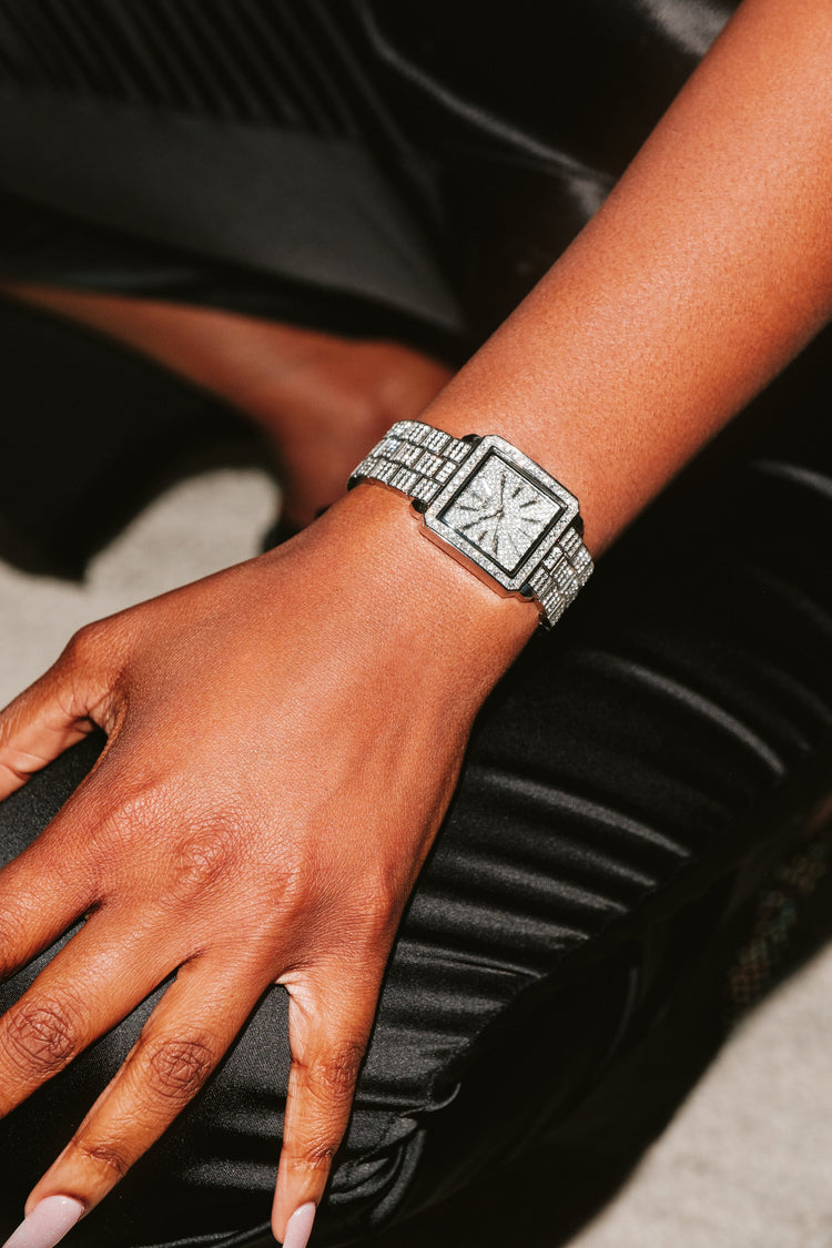 A silver-toned square watch with a diamond-encrusted face and bracelet, worn on a person's wrist while they rest their hand on black pleated fabric.