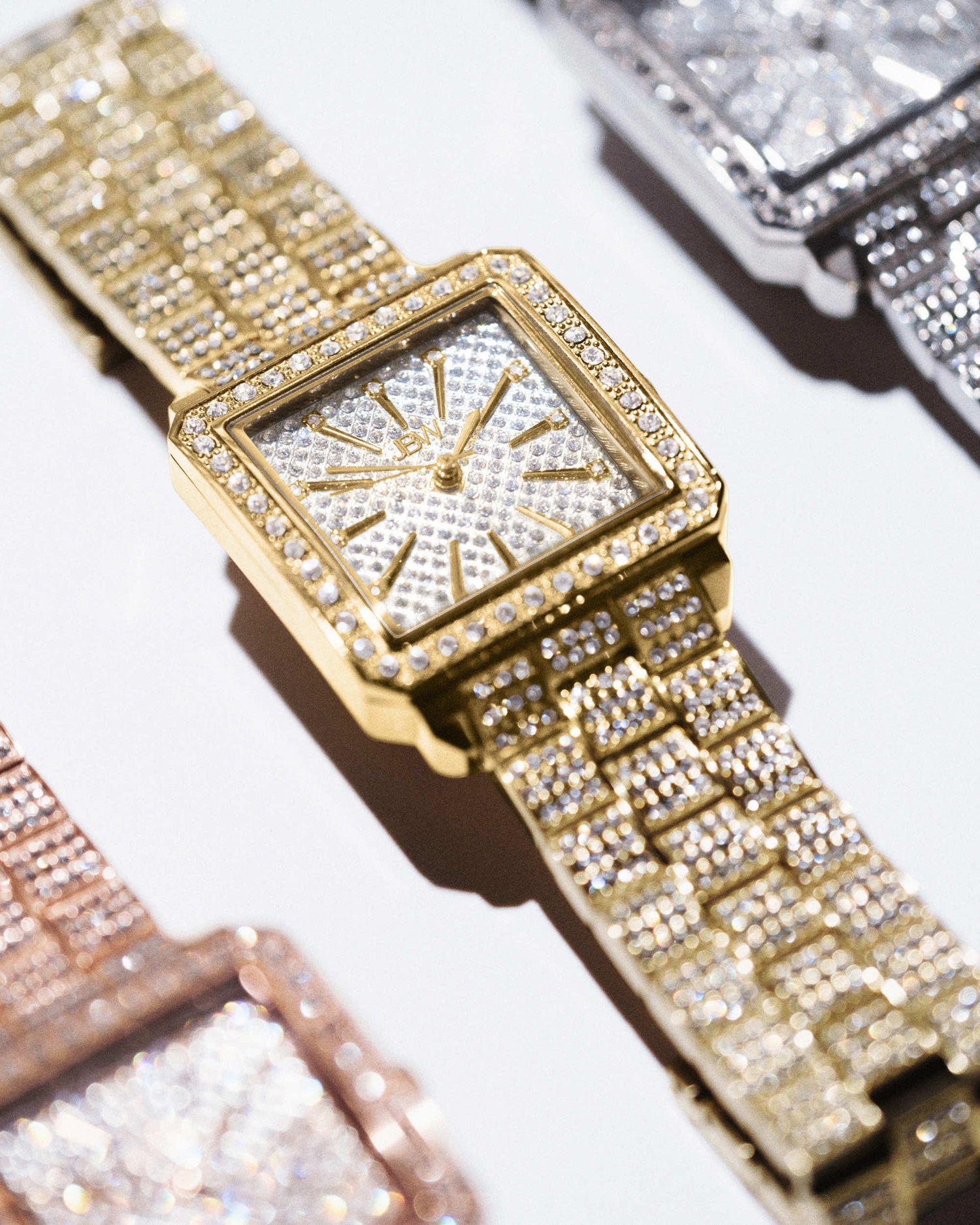 The JBW Cristal 28 | J6386A, a gold-plated stainless steel square wristwatch with diamond accents, rests on a white surface alongside other jeweled JBW watches in silver and rose gold tones.