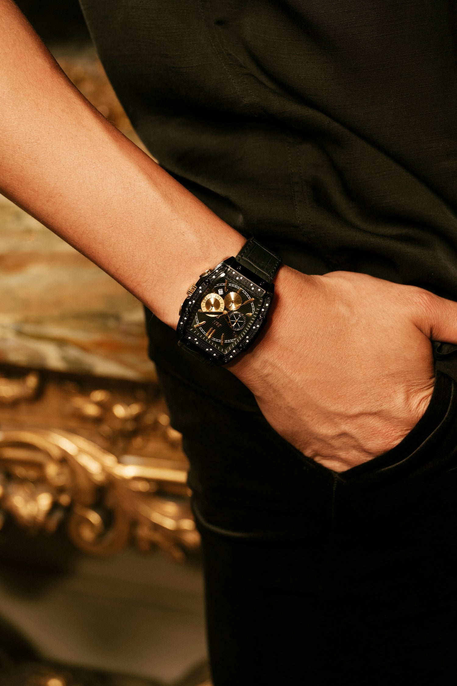 A close-up lifestyle shot of a person's wrist wearing a black sports watch with a gold-toned dial and crystal-encrusted bezel, paired with a black shirt and trousers.