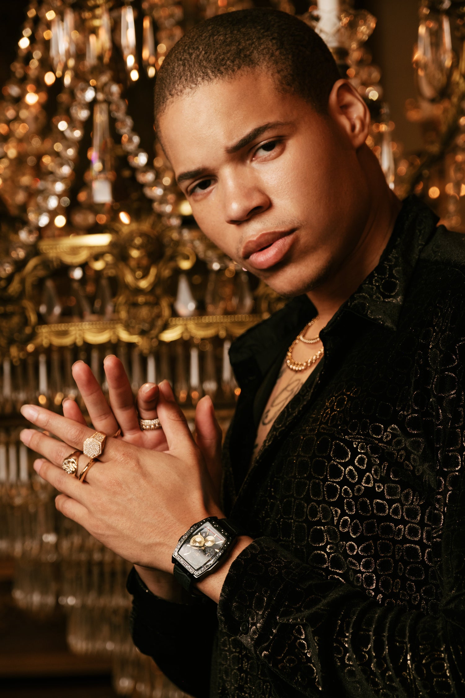 A man in a black patterned shirt and gold jewelry posing in front of a large, ornate gold chandelier.