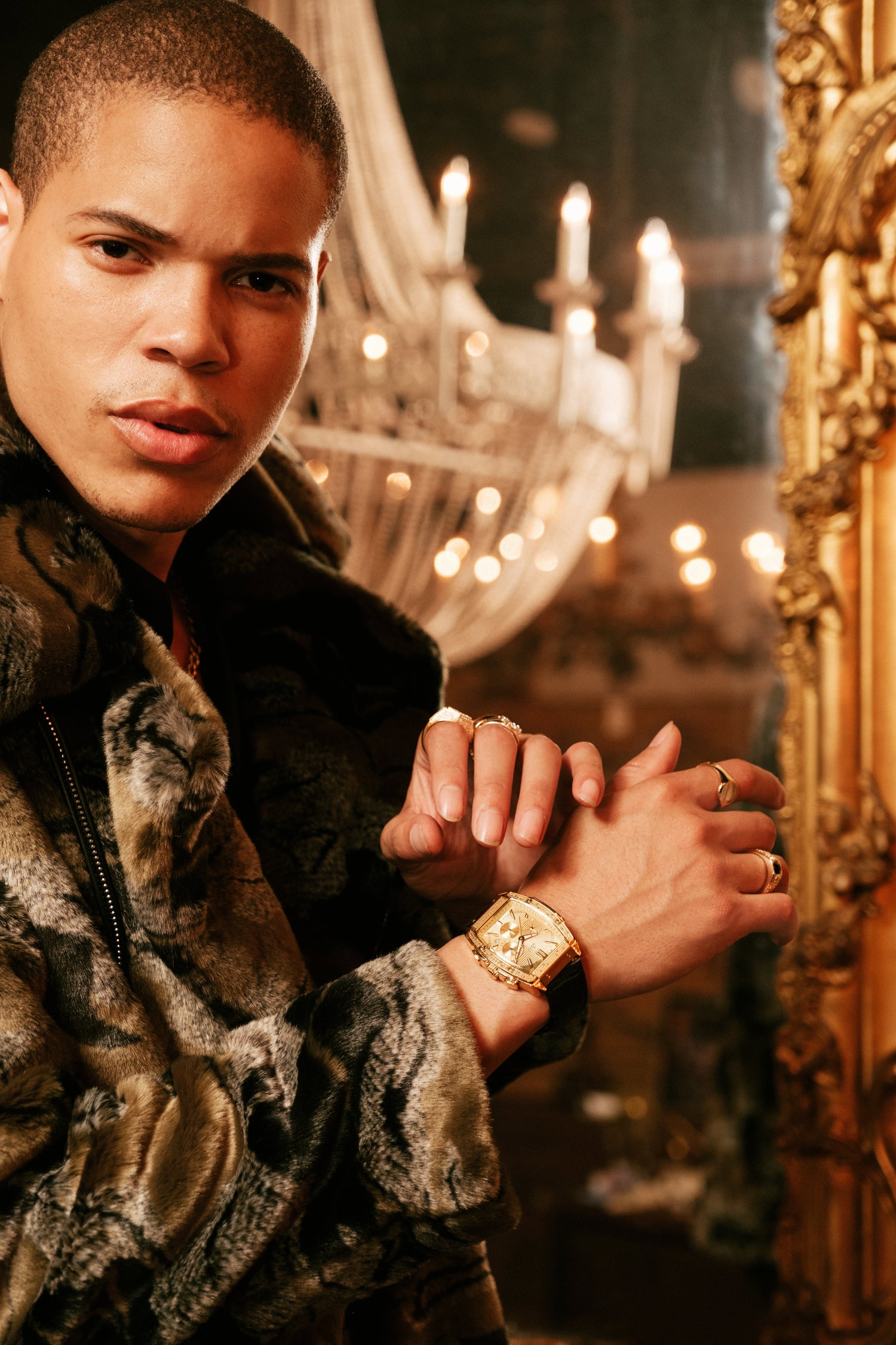A man in a brown and black patterned fur jacket poses in an ornate room with a large gold mirror and a chandelier in the background, wearing a gold watch and rings.