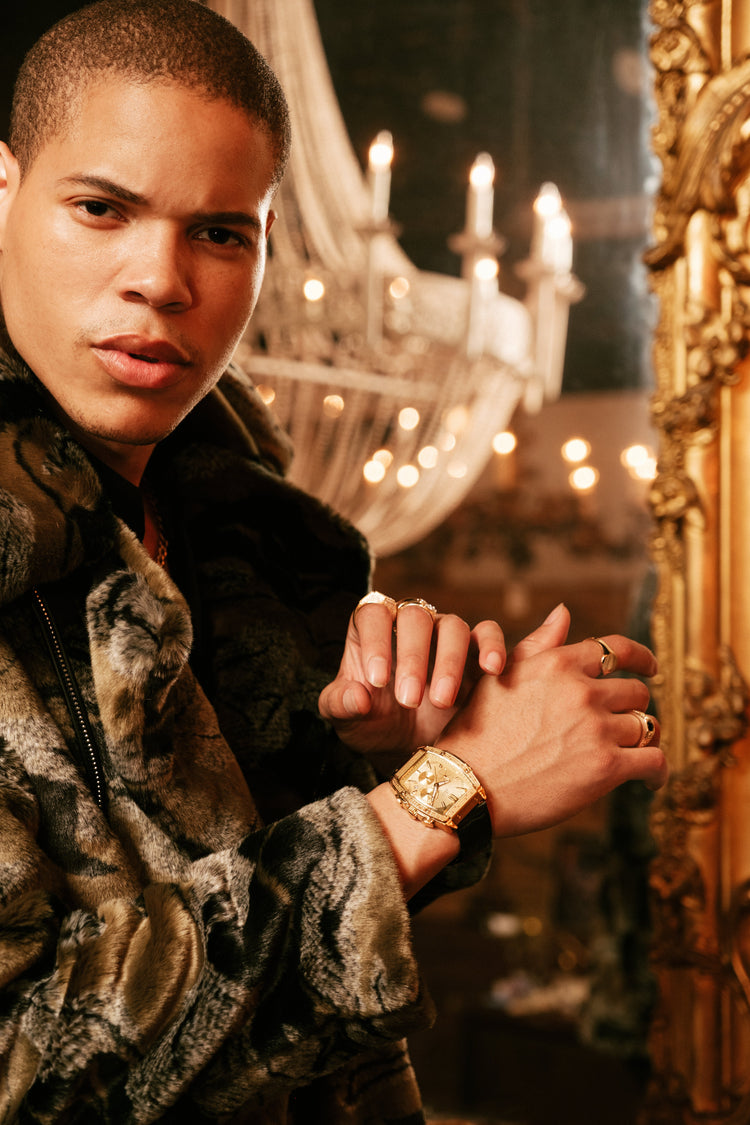 A man in a brown and black patterned fur jacket poses in an ornate room with a large gold mirror and a chandelier in the background, wearing a gold watch and rings.