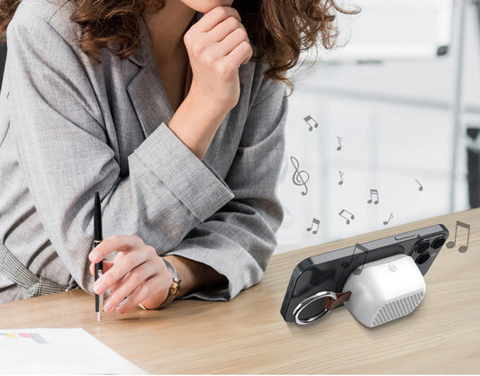 A woman in a gray blazer sits at a wooden desk, looking down at a black smartphone case with a built-in white speaker and ring light. Musical note graphics float around the device, suggesting sound output, in a bright, modern office setting.