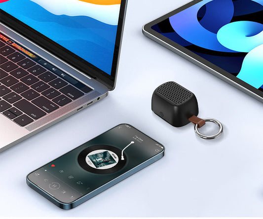A small black portable speaker with a key ring attached, shown on a clean light gray surface alongside a laptop, tablet, and smartphone displaying media.