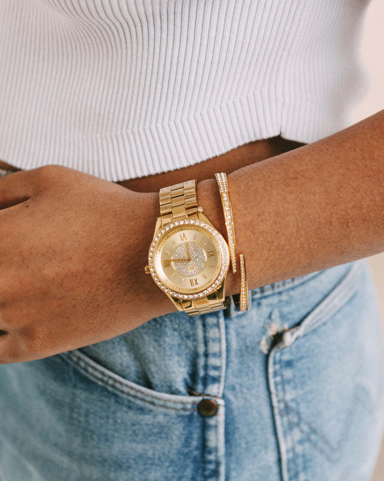 Wearing the JBW Mondrian | J6303B 18K gold-plated wristwatch and a gold bracelet, a person stands with one hand in the pocket of light blue jeans, dressed in a white ribbed top.