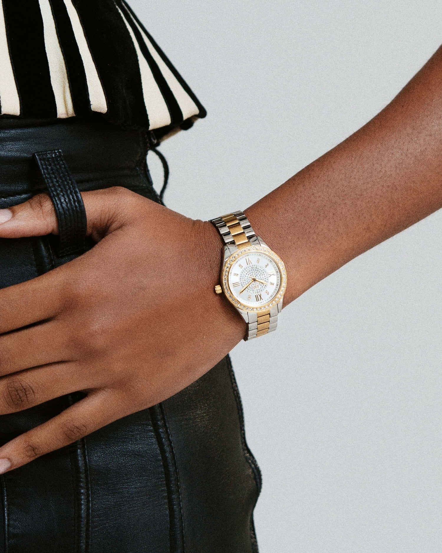 A person in black leather pants and a striped black-and-white top rests their hand on their hip, wearing the JBW Mondrian | J6303G gold-plated stainless steel wristwatch with a white face.