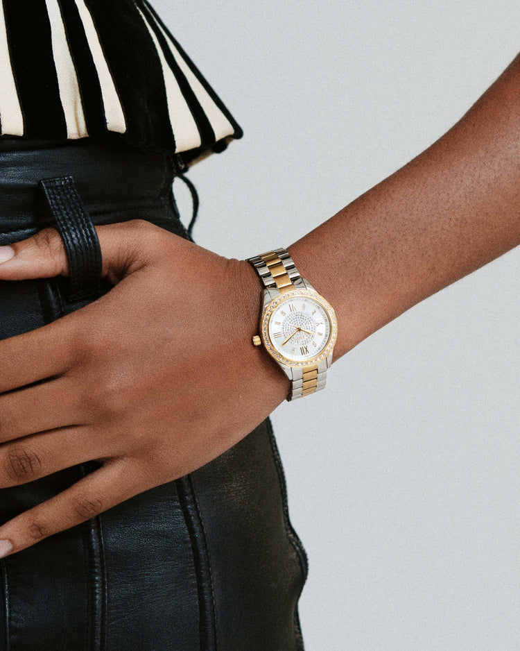 A person in black leather pants and a striped black-and-white top rests their hand on their hip, wearing the JBW Mondrian | J6303G gold-plated stainless steel wristwatch with a white face.