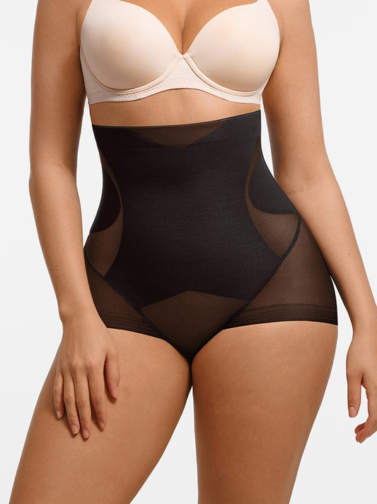 A model wearing high-waisted black shapewear briefs with sheer mesh side panels, paired with a beige bra, against a plain light gray studio background.