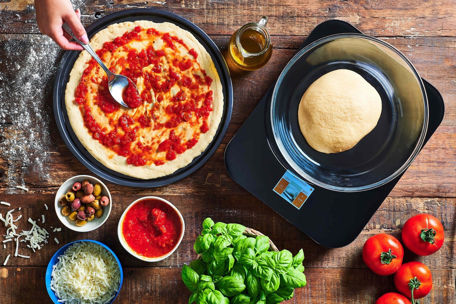 Person preparing a pizza with ingredients on a wooden table