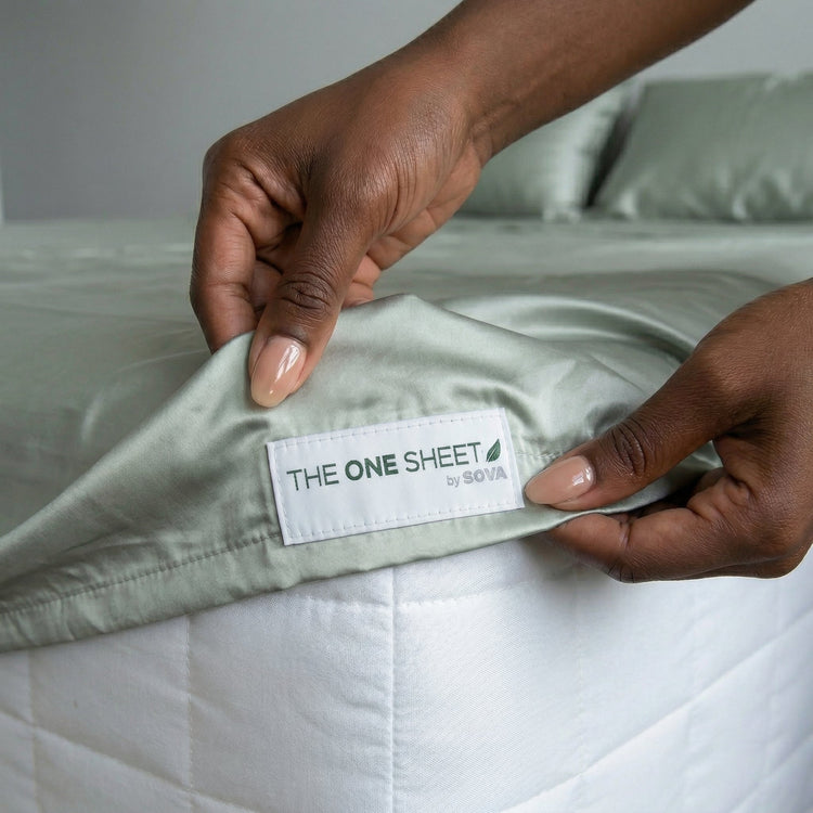 Person holding a folded sheet with 'The One Sheet' label on a bed.