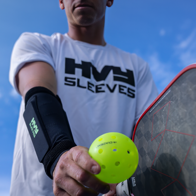 Person holding a pickleball and paddle with 'HVY SLEEVES' branding against a blue sky.
